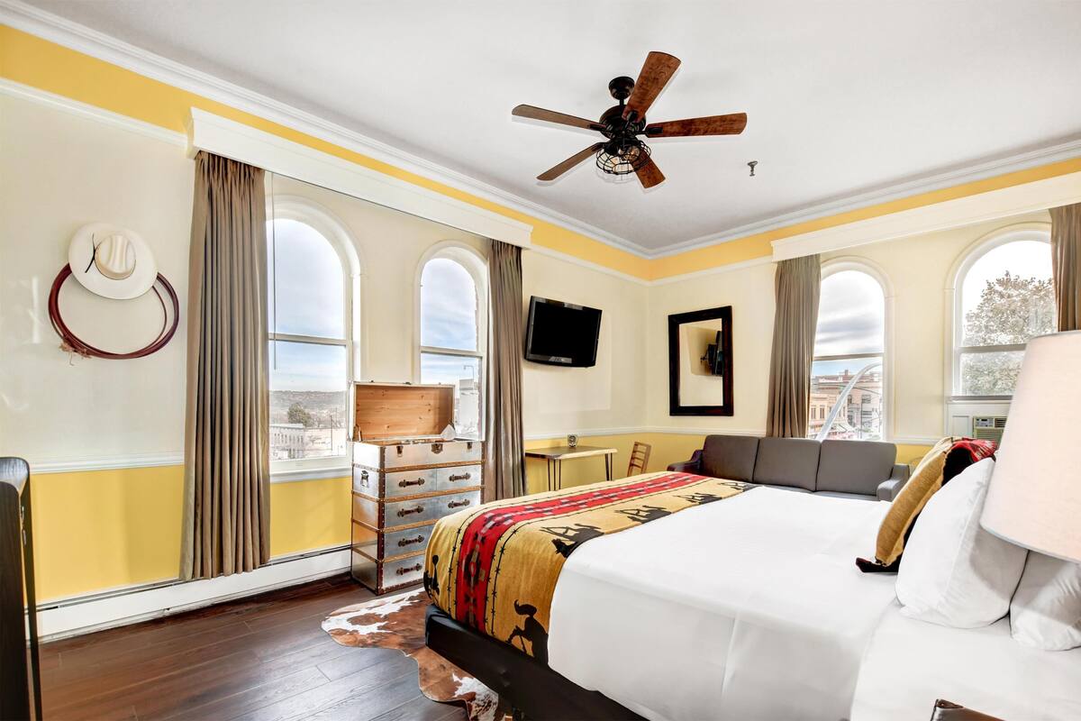 Habitación de hotel con una cama grande, ventilador de techo, escritorio con silla, televisor y ventanas amplias con cortinas.