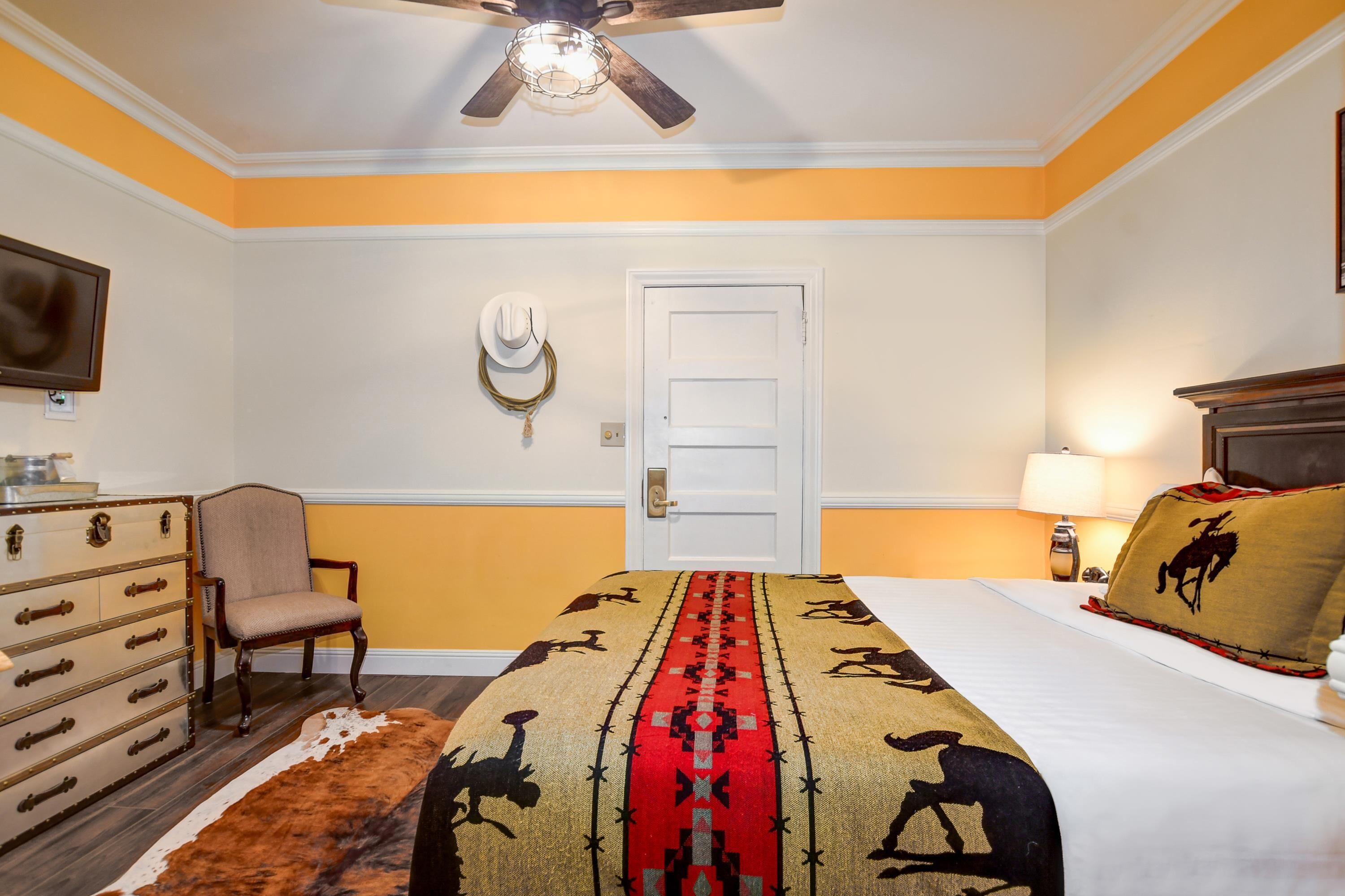 A hotel room with a bed, a dresser, a chair, a ceiling fan, a door, and a lamp.