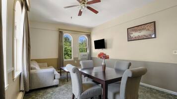 Uma sala de jantar com mesa, cadeiras, ventilador de teto, televisão e janela com vista para árvores.
