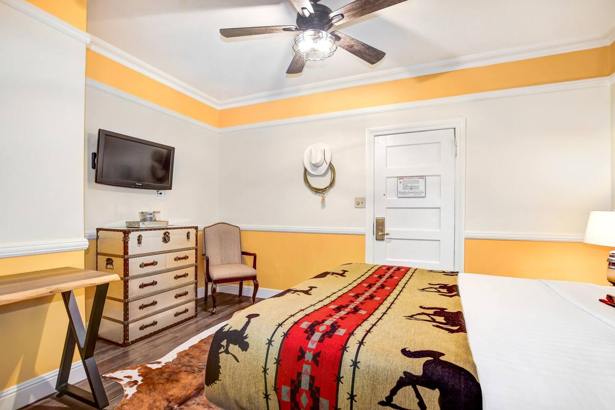 Habitación de hotel con cama, escritorio, silla, televisión, puerta y ventilador de techo.