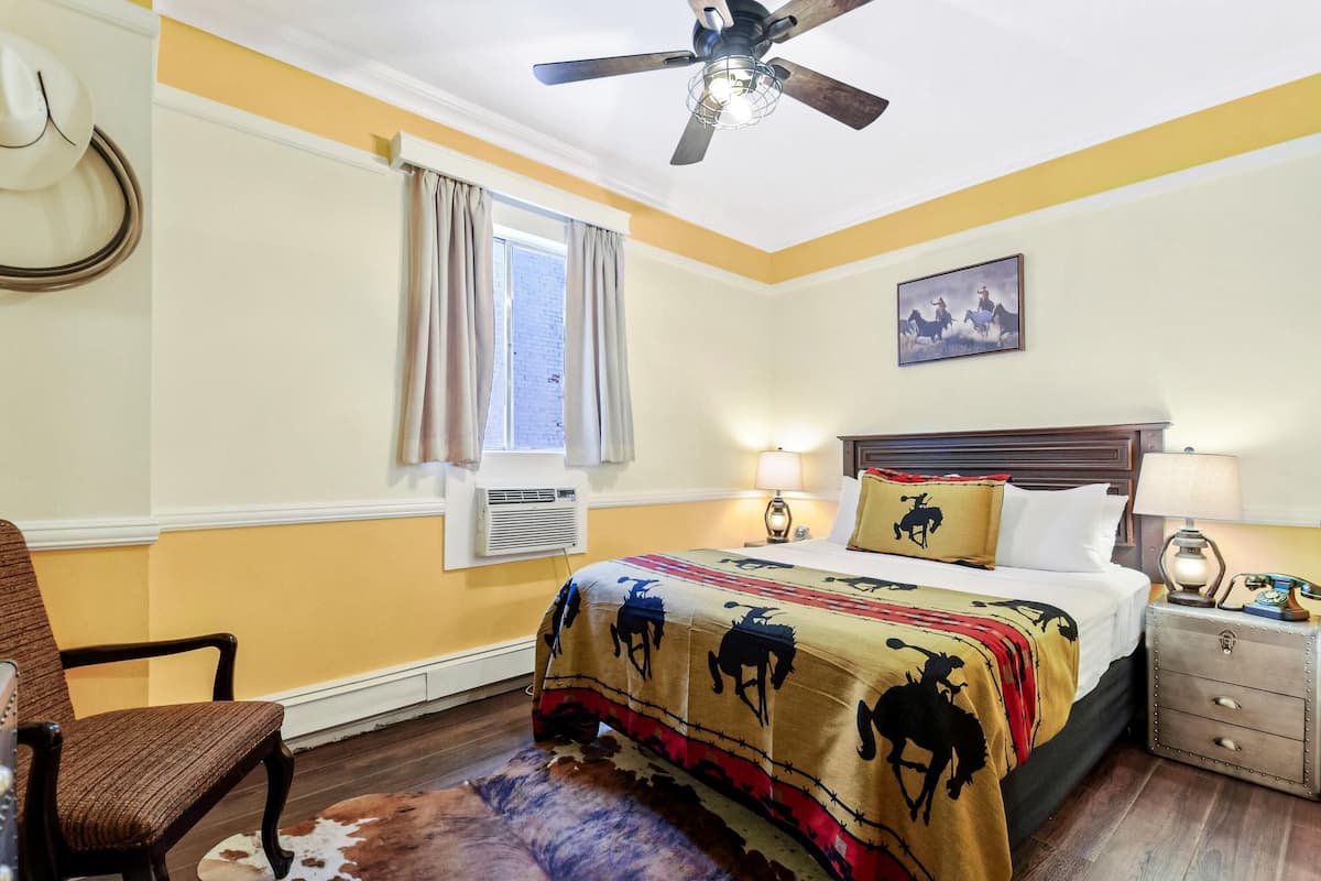 Una habitación de hotel con cama, una silla, una mesita de noche, ventilador de techo, ventana con cortinas y aire acondicionado de pared.