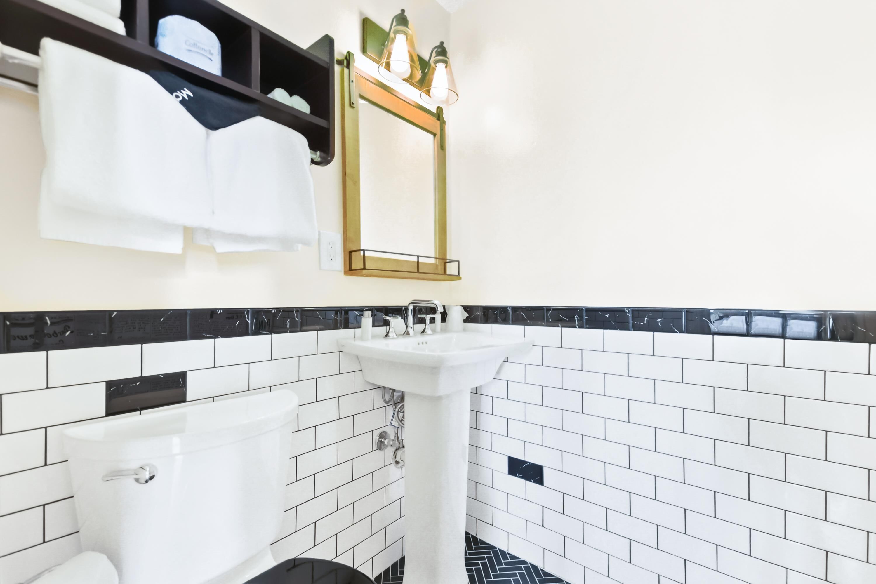 A bathroom with a white pedestal sink, a white bathtub, black and white tiled walls, and a mirror with three lights.