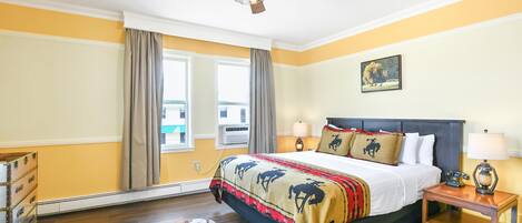 Een slaapkamer met een bed, nachtkastje, plafondventilator, raam met gordijnen, een wandairconditioner en een schilderij.