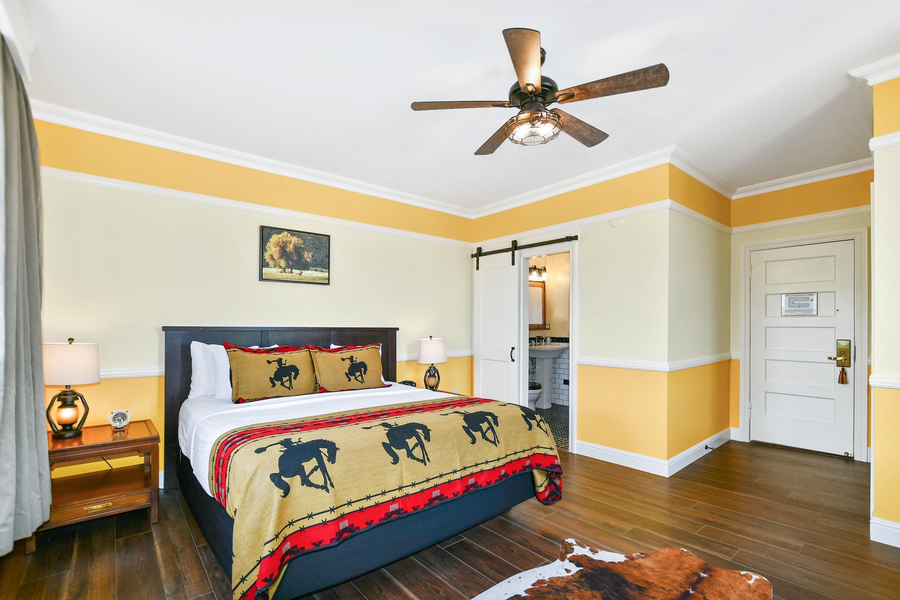 Une chambre d’hôtel avec un lit, un ventilateur de plafond, une table de chevet, une lampe, une porte et une salle de bain visible à travers une porte ouverte.