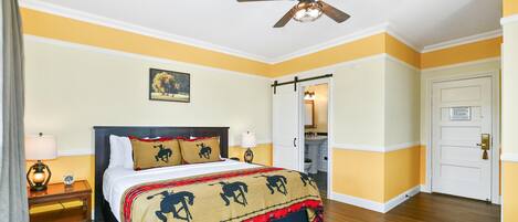 Une chambre d’hôtel avec un lit, un ventilateur de plafond, une table de chevet, une lampe, une porte et une salle de bain visible à travers une porte ouverte.
