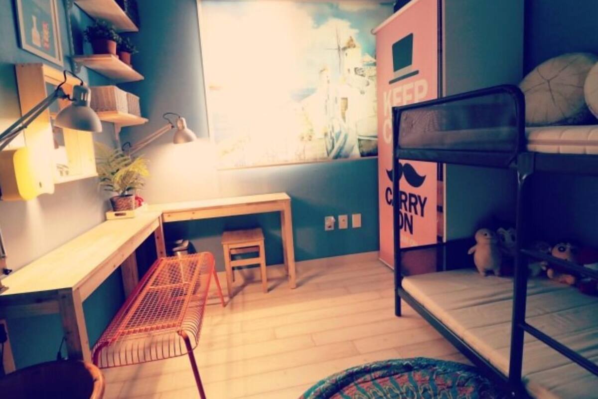 A compact bedroom with a bunk bed, a desk with a chair, a small stool, and a shelf with plants.