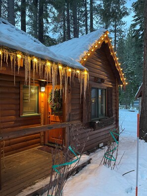 Una cabaña de madera con borbotones de hielo colgando del techo, un porche con un banco y un terreno cubierto de nieve.