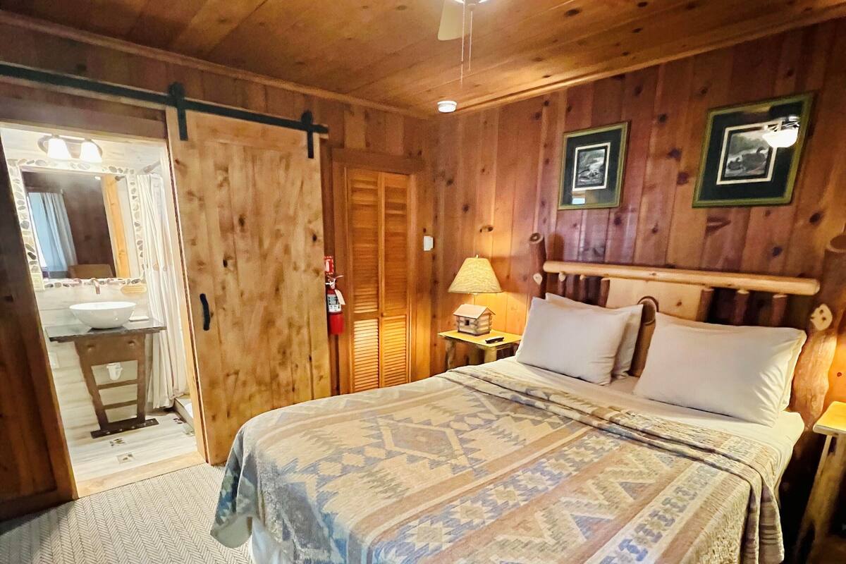 Un dormitori de cabana de fusta amb un llit, una tauleta de nit, una làmpada, un ventilador de sostre i una porta que condueix a un bany.