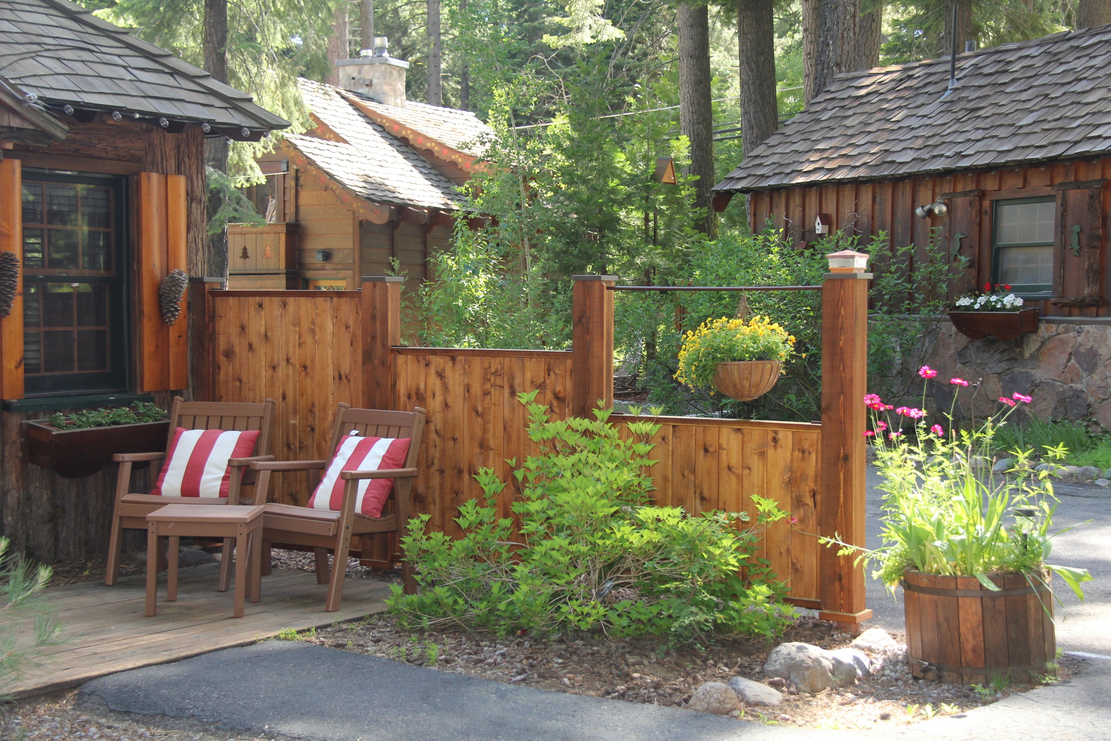 A wooden cabin with a deck, two chairs with striped cushions, and a garden area with plants and flowers.