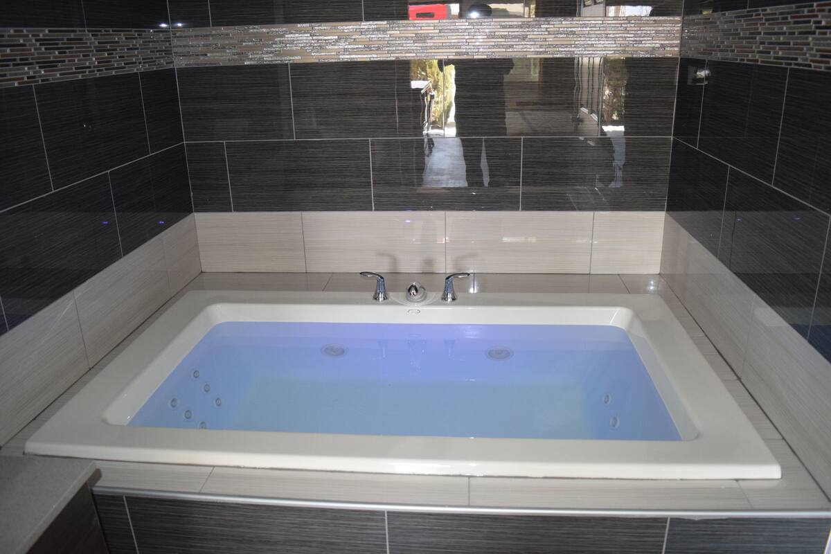 A rectangular bathtub with two faucets, surrounded by dark tiles with a reflective strip above.