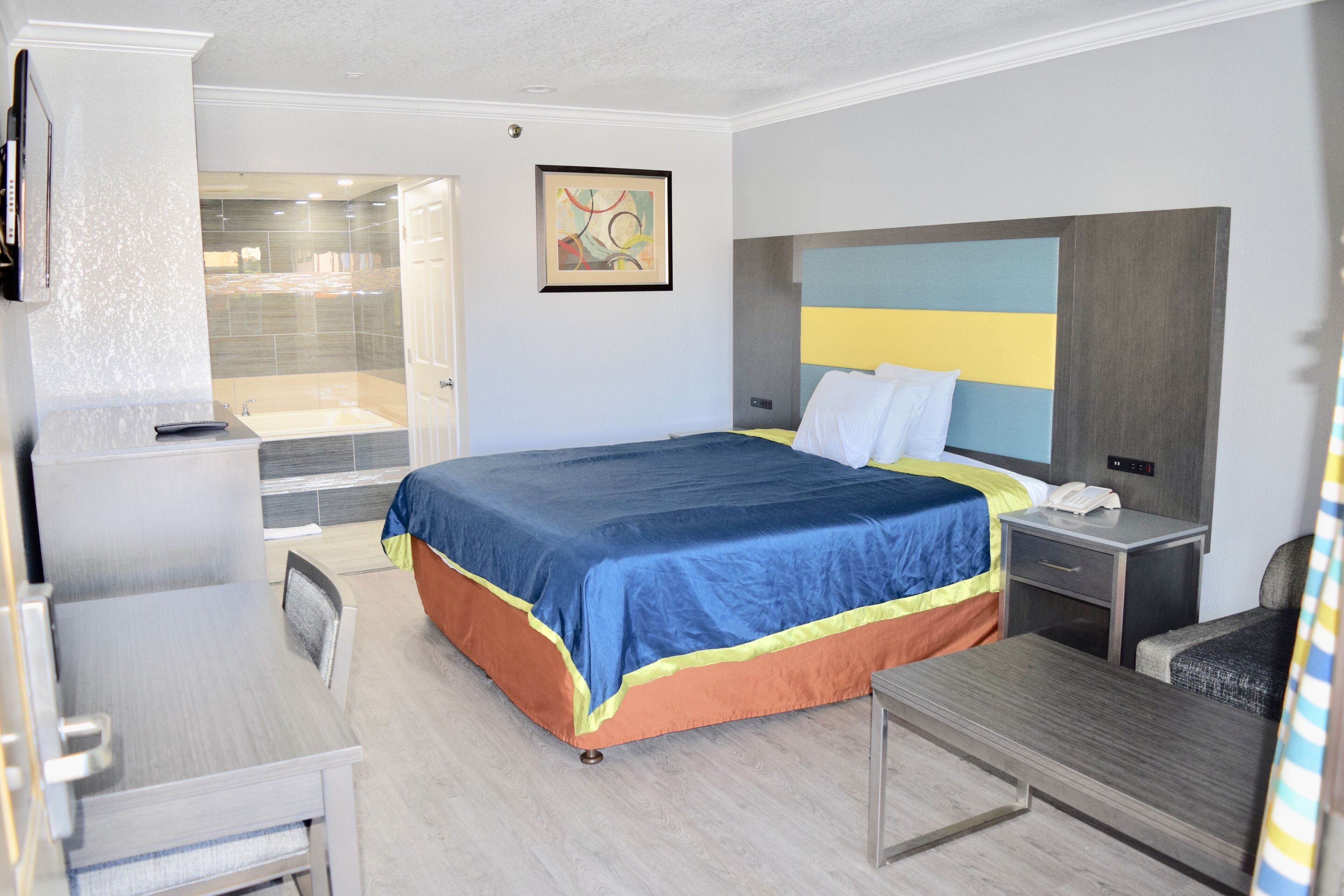 Habitación de hotel con cama, mesita de noche, silla, escritorio y baño con ducha y puerta de vidrio.
