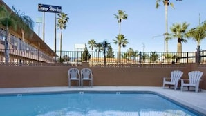 Uma piscina com duas cadeiras brancas, uma cerca e palmeiras ao fundo.