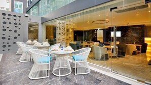 Un espacio al aire libre con sillas y mesas de mimbre blanco, un muro de cristal que separa el área de comedor de la calle, y una fachada de edificio moderna al fondo.