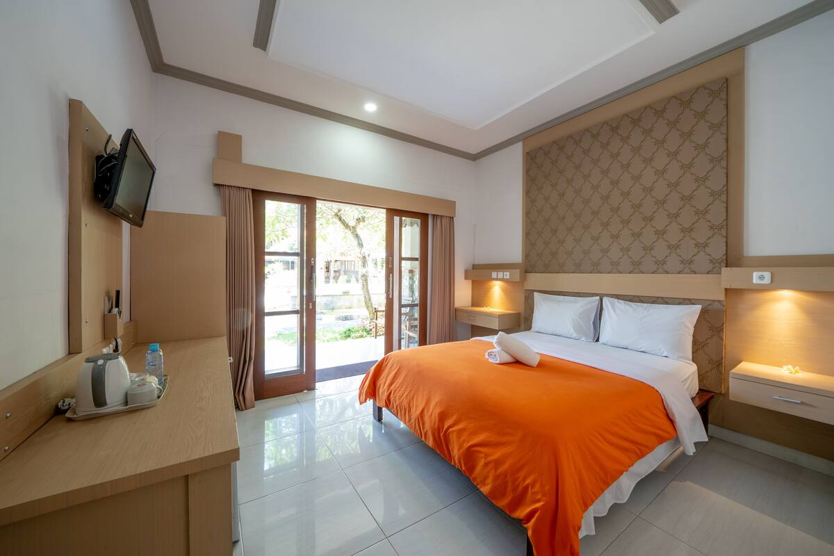 A hotel room with a bed, a desk with a kettle, a TV, and a sliding glass door to the outside.