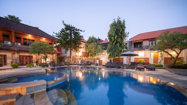 A swimming pool with lounge chairs and umbrellas, surrounded by buildings with red-tiled roofs and outdoor lighting. - Bendesa Accommodation (Kuta)