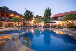 A swimming pool with lounge chairs and umbrellas, surrounded by buildings with red-tiled roofs and outdoor lighting. - Bendesa Accommodation (Kuta)