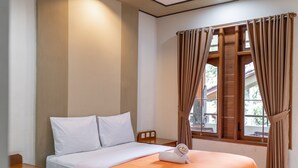A neatly made bed with white and orange bedding, a wooden nightstand, and a window with brown curtains. - Bendesa Accommodation (Kuta)