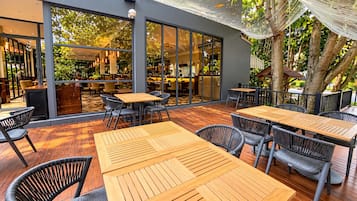 An outdoor seating area with wooden tables and chairs, large windows, and a view of the interior.