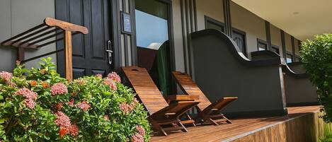 A modern outdoor seating area with wooden loungers and a small table, surrounded by lush greenery and a dark-colored building facade.