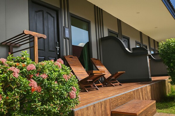 Un espacio exterior moderno con sillones de madera y una pequeña mesa, rodeado de abundante vegetación y una fachada de edificio de color oscuro.