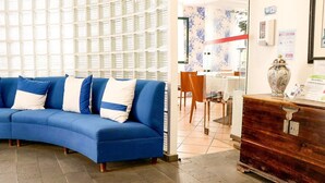 A blue sofa with white cushions in a lobby area with a tiled floor and a wooden cabinet with a vase on top. - Hotel Amparo (Machico)