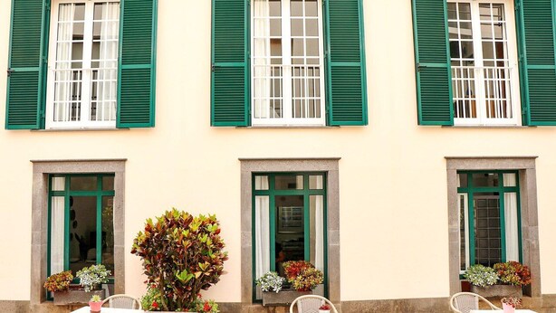 A building with three windows, each with green shutters and white frames. There are outdoor tables and chairs, and potted plants near the windows. - Hotel Amparo (Machico)