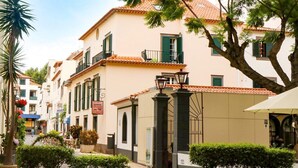 A building with a tiled roof, a balcony, and a street with shops and greenery. - Hotel Amparo (Machico)