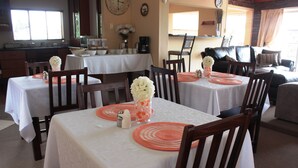 A dining area with multiple tables set for service, a kitchen in the background, and a living area with a sofa and chairs. - Sunnyridge Lodge (East London)