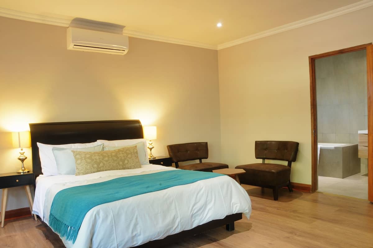 Quarto de hotel com uma cama grande, duas mesas de cabeceira com luminárias, um painel de madeira na cabeceira, um banquinho e uma porta que dá para um banheiro.
