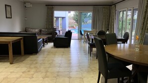 A spacious room with a sofa, chairs, a dining table, and a view of a swimming pool through large windows. - Ilanda Guest House (Mbombela)