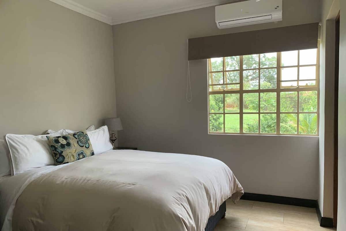 Quarto com cama grande, janela com vista para o verde, ar condicionado e mesa de cabeceira com luminária.