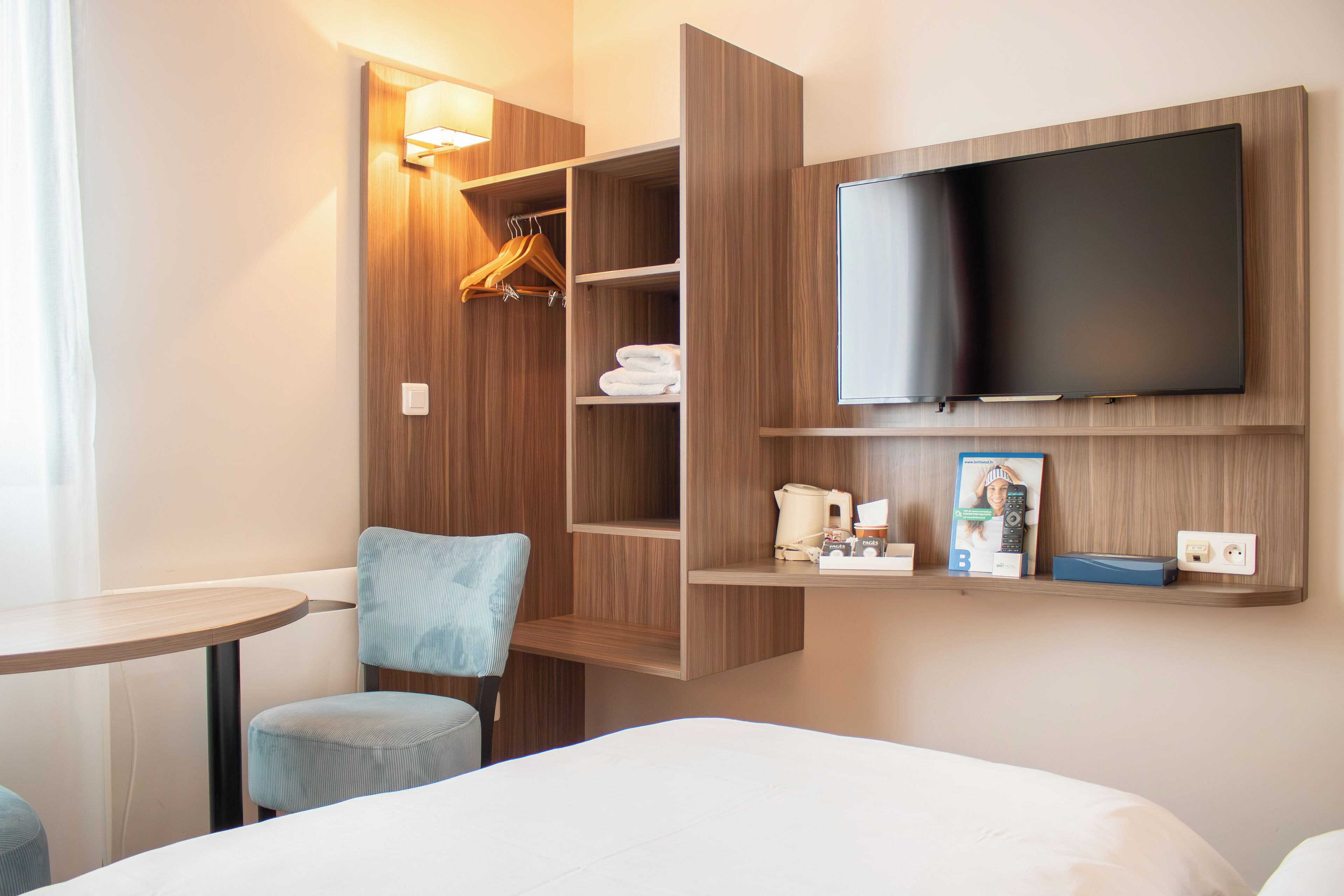 Une chambre d’hôtel comprenant un lit, une chaise, une table, un téléviseur fixé au mur, une armoire avec des étagères et une petite étagère sur laquelle sont posés divers objets.