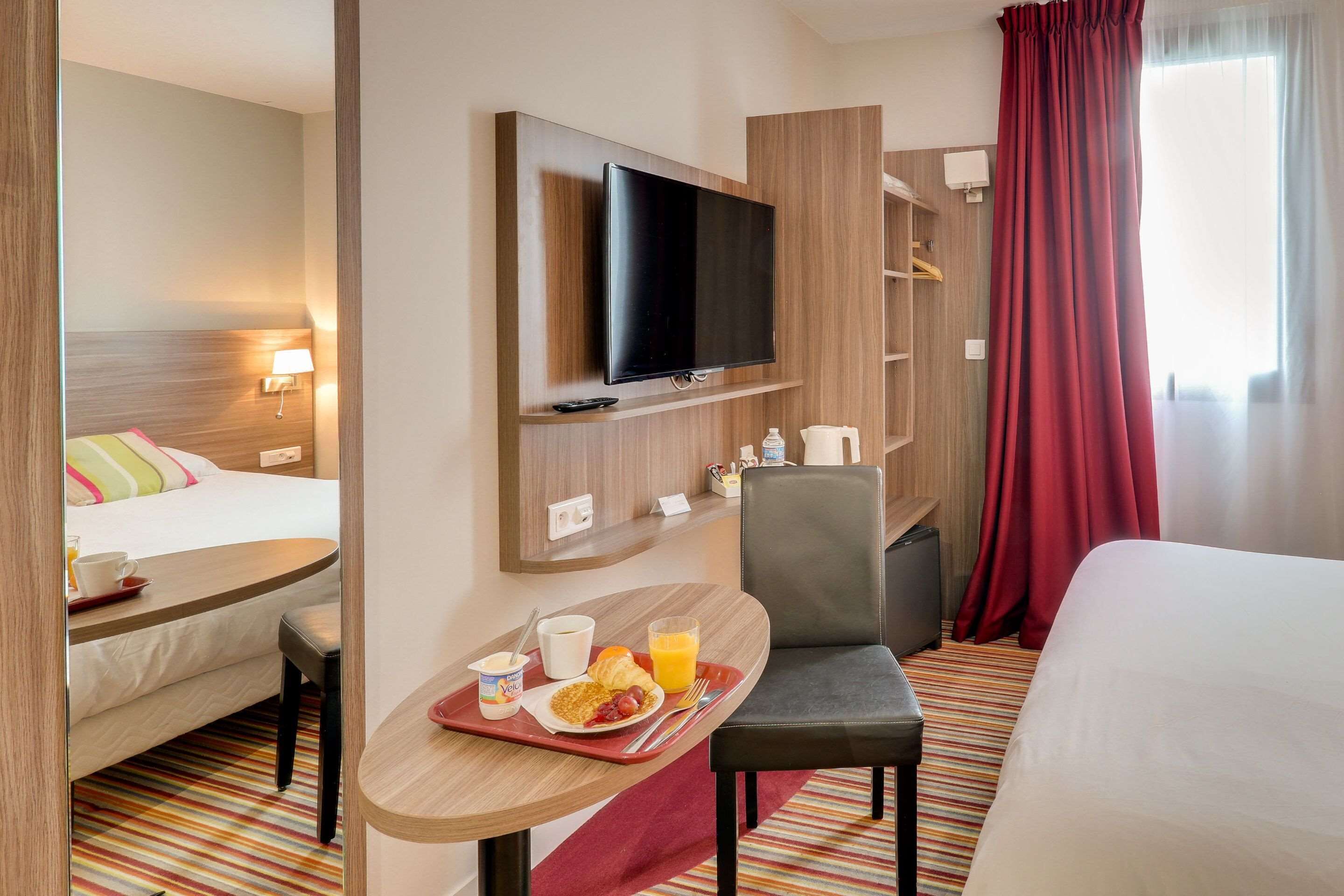 Habitación de hotel con cama, televisor montado en la pared, una mesita con cosas para el desayuno, una silla y ventana con cortinas.