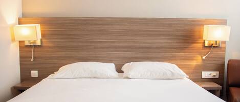 Camera d'hotel con una testiera in legno, due lampade sui comodini, un letto con lenzuola bianche e una poltrona in pelle marrone.