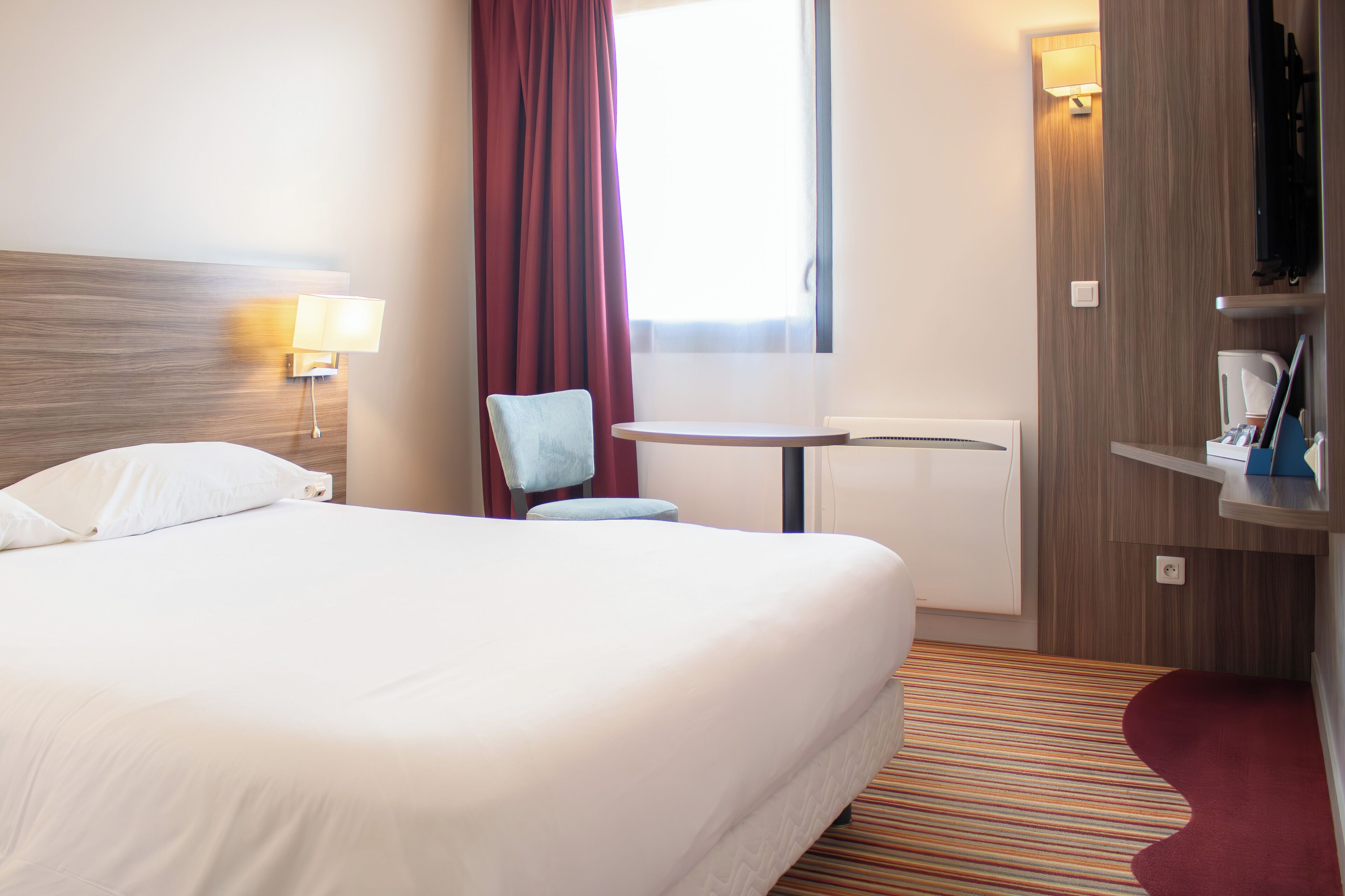 Une chambre d’hôtel avec un grand lit, une chaise, une petite table, une télévision et une fenêtre avec des rideaux.