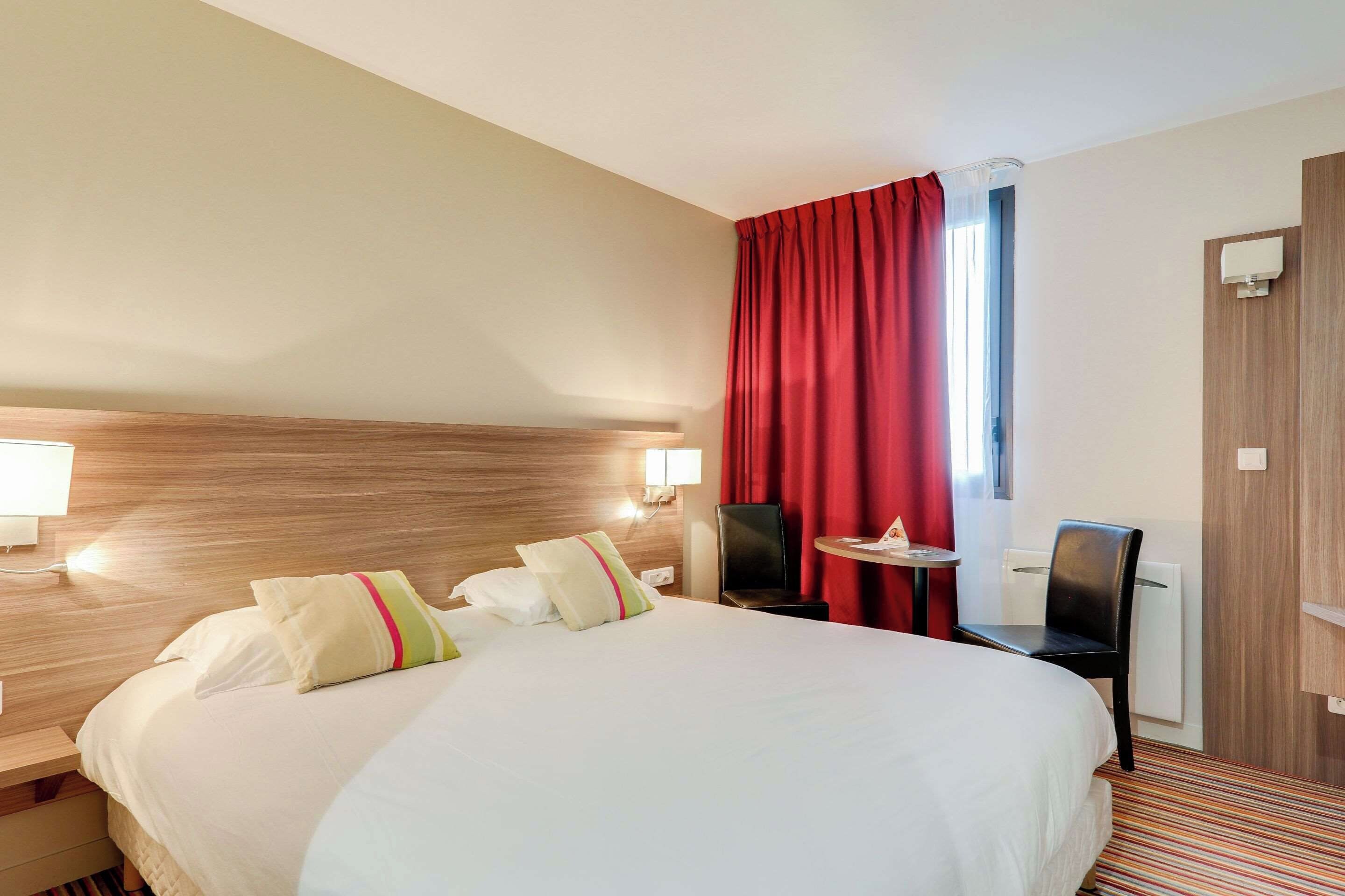 A hotel room with a large bed, two bedside lamps, a small table, a chair, and a window with red curtains.