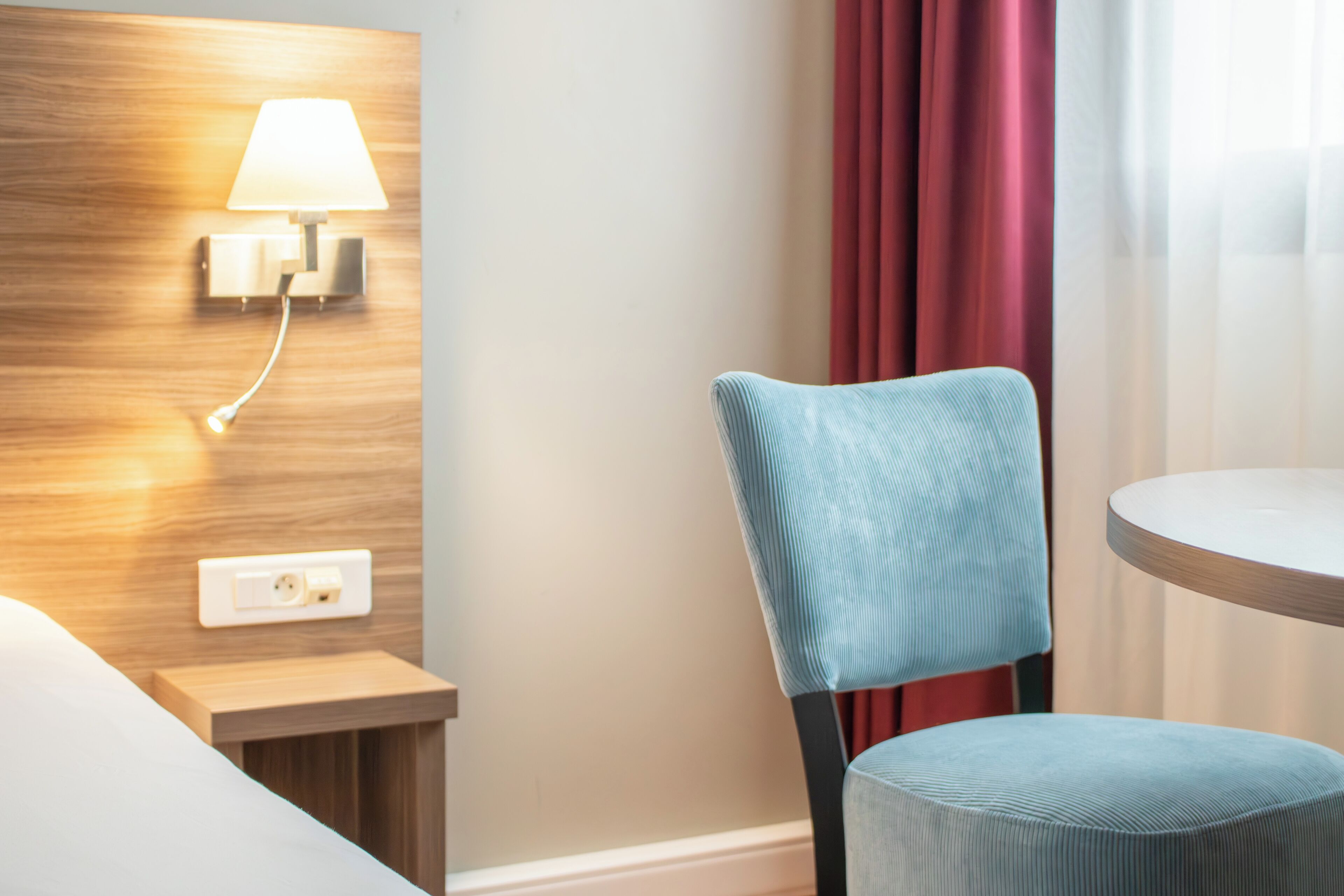 Ein Hotelzimmer mit einem hölzernen Kopfteil, einer Wandlampe, einem blau gepolsterten Sessel und einem runden Tisch.