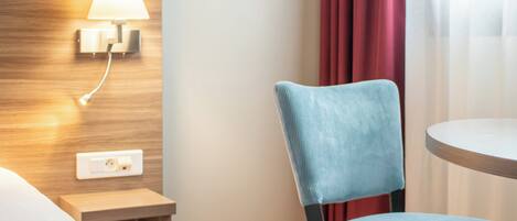 A hotel room with a wooden headboard, a wall-mounted lamp, a blue upholstered chair, and a round table.