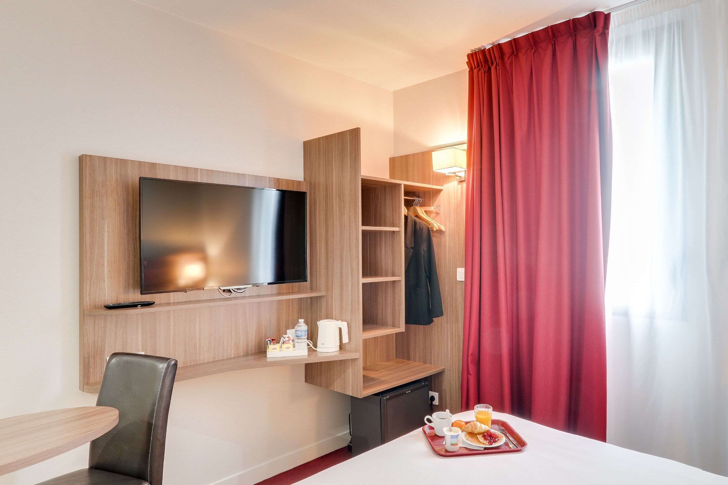 A hotel room with a flat-screen TV mounted on the wall, a small kitchenette, a dining area with a table and chair, and a breakfast tray on the table.