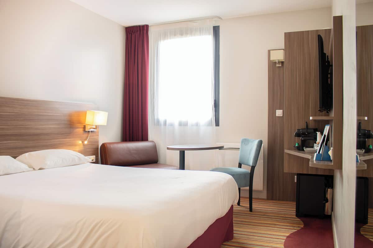 A hotel room with a large bed, a desk with a chair, a small table, and a TV on a stand.