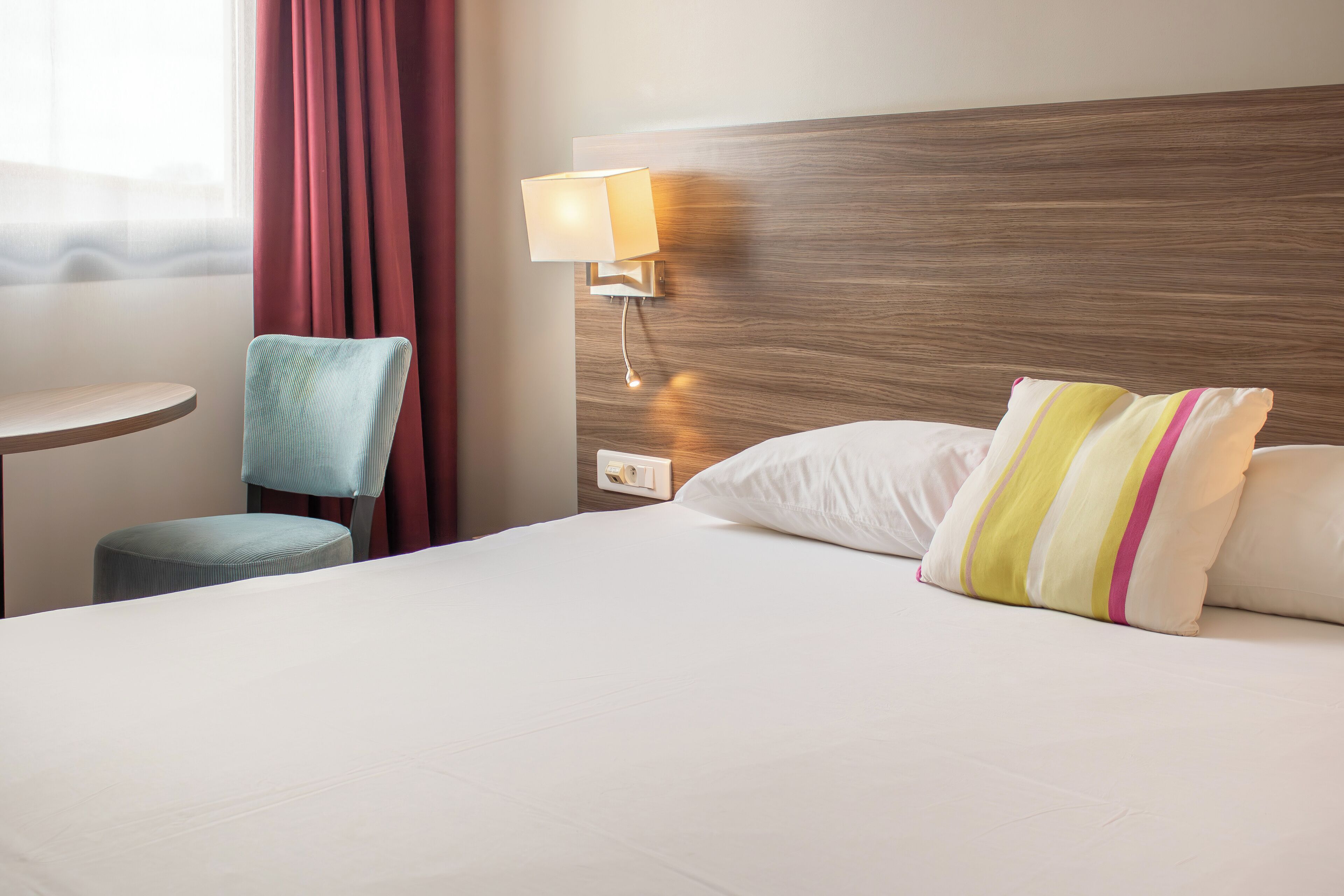 Una habitación de hotel con cama, una silla, mesita de noche, lámpara y ventana con cortinas.