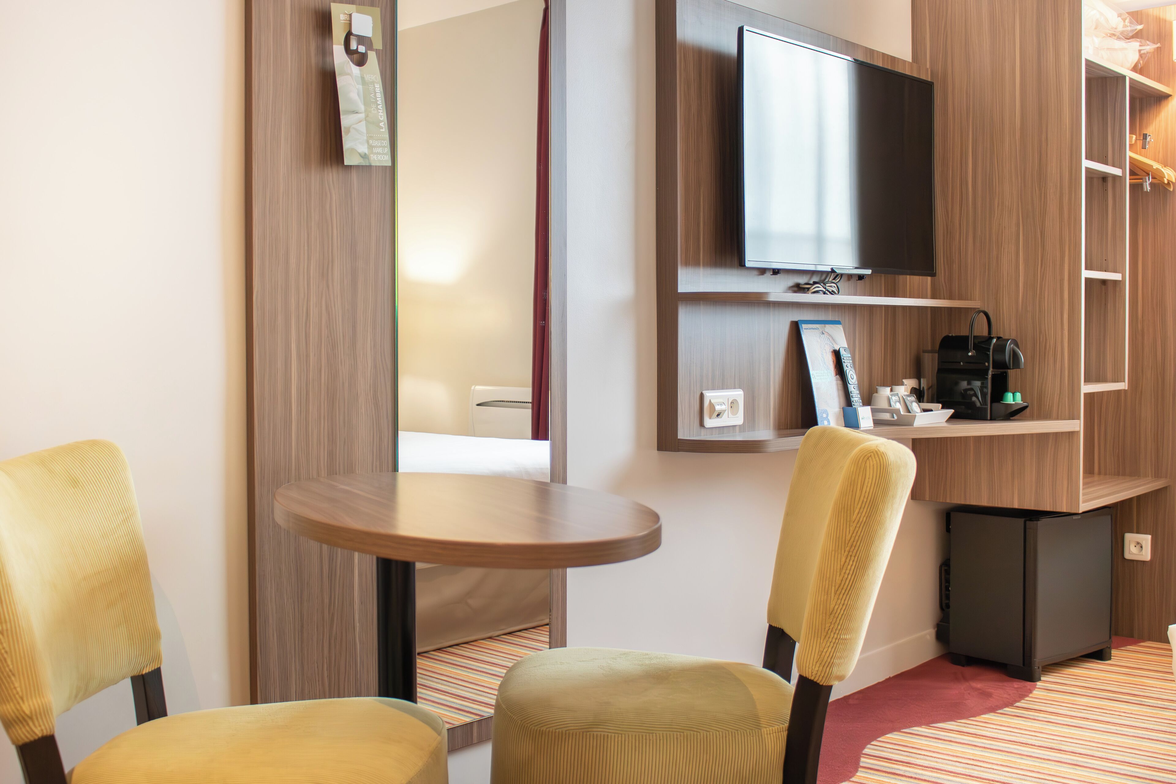 A hotel room with a flat-screen TV mounted on the wall, a small table, two chairs, a compact refrigerator, and a wooden shelving unit with a few items on it.