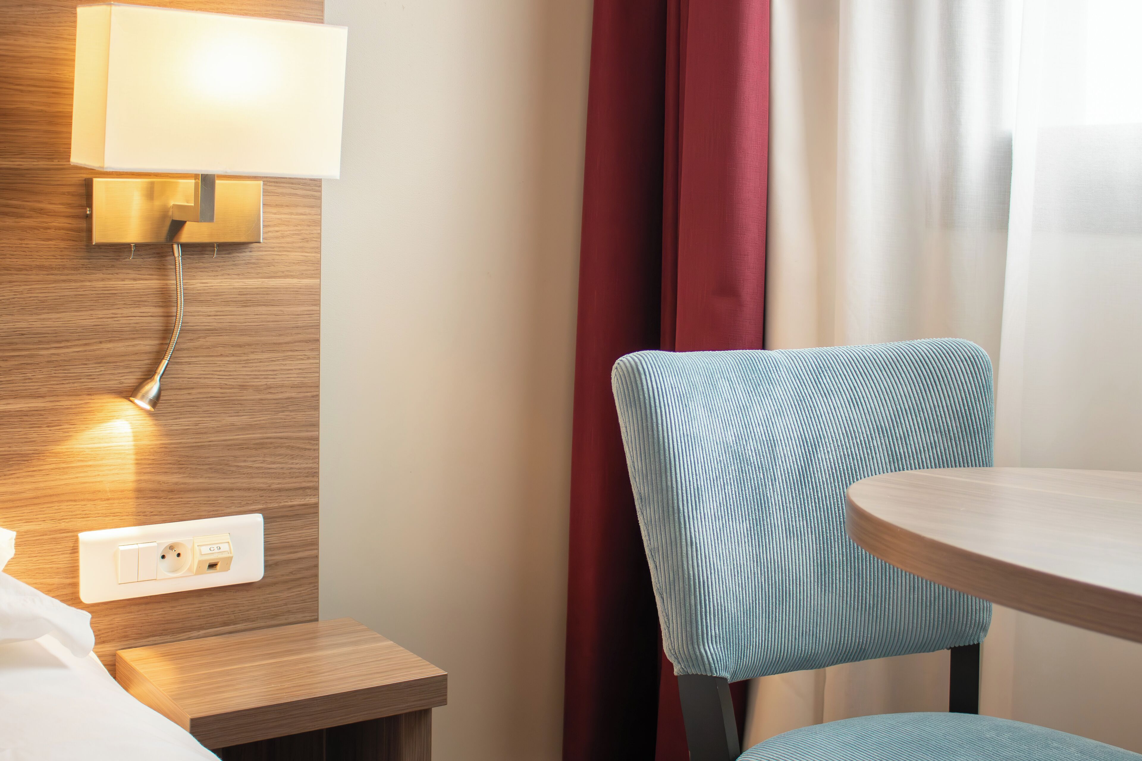 A hotel room with a wooden headboard, a bedside table, a lamp, a chair, and a round table.