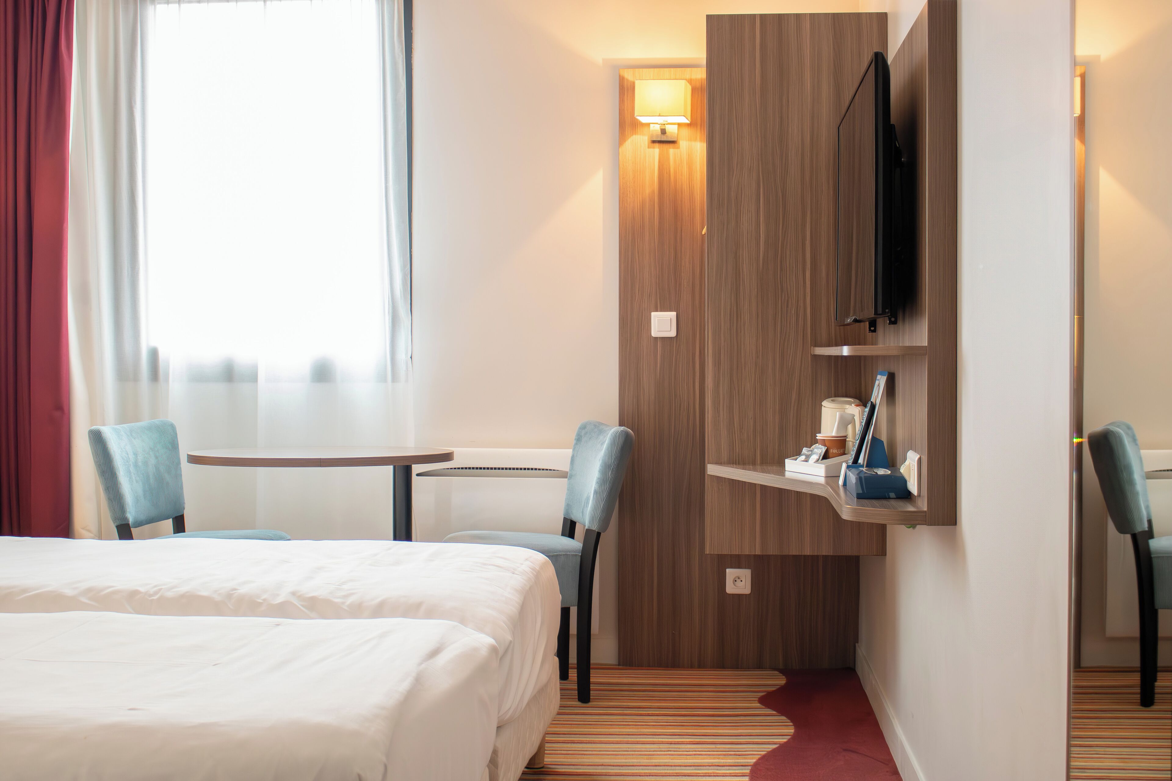 Hotelkamer met een bed, twee stoelen, een kleine tafel, een aan de muur bevestigde televisie en een lamp.
