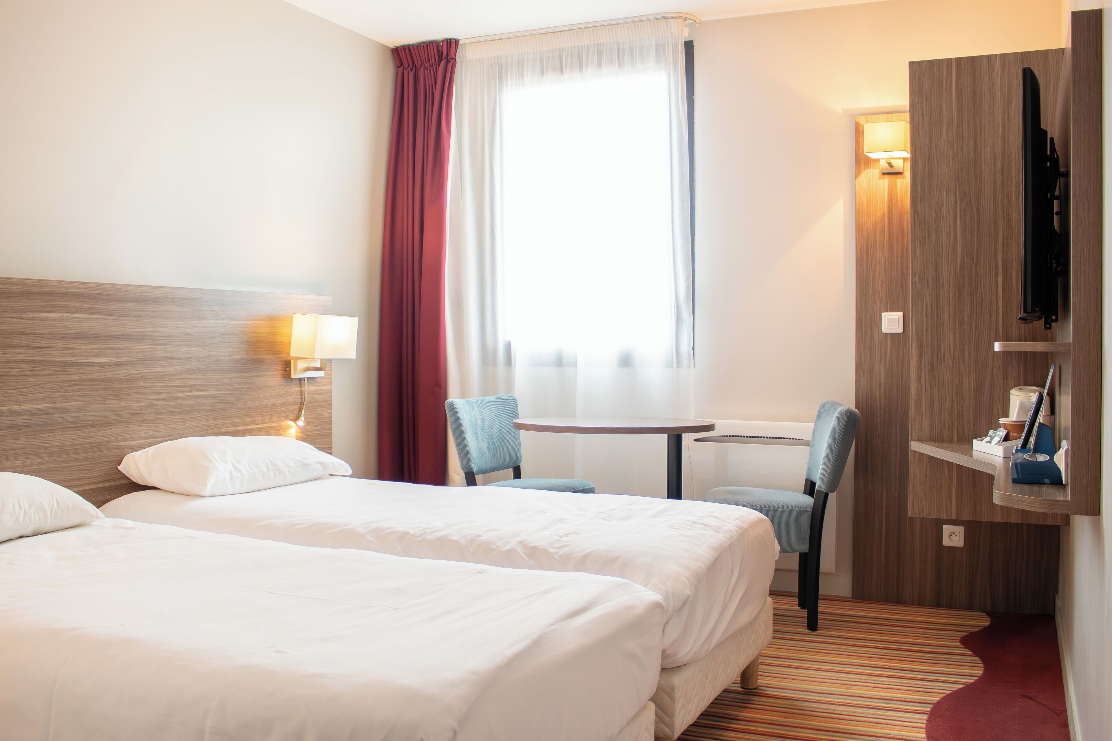 A hotel room with two beds, a desk, a chair, a TV, and a window with curtains.
