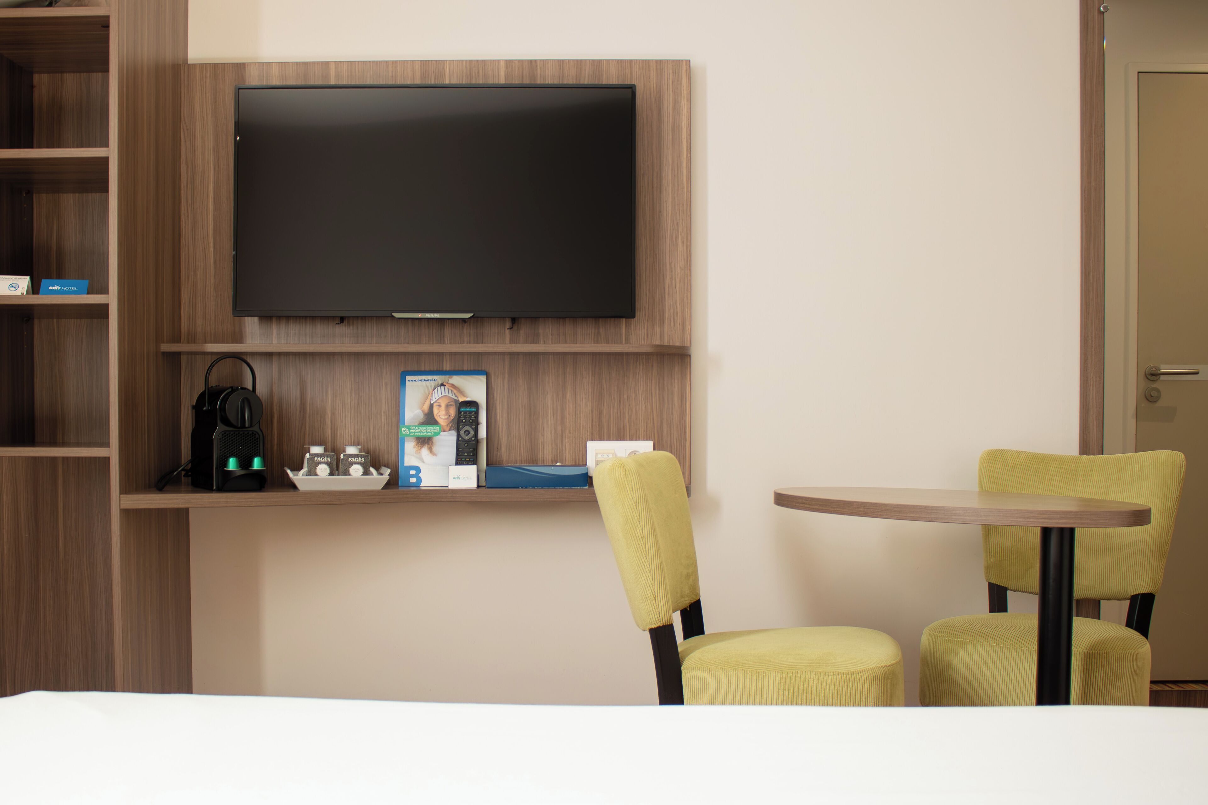 A hotel room with a flat-screen TV mounted on the wall, a coffee maker, and a small table with two yellow chairs.