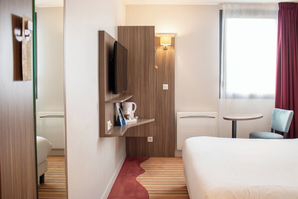 Habitación de hotel con cama, televisor montado en la pared, una mesita con una silla y una ventana con cortinas.