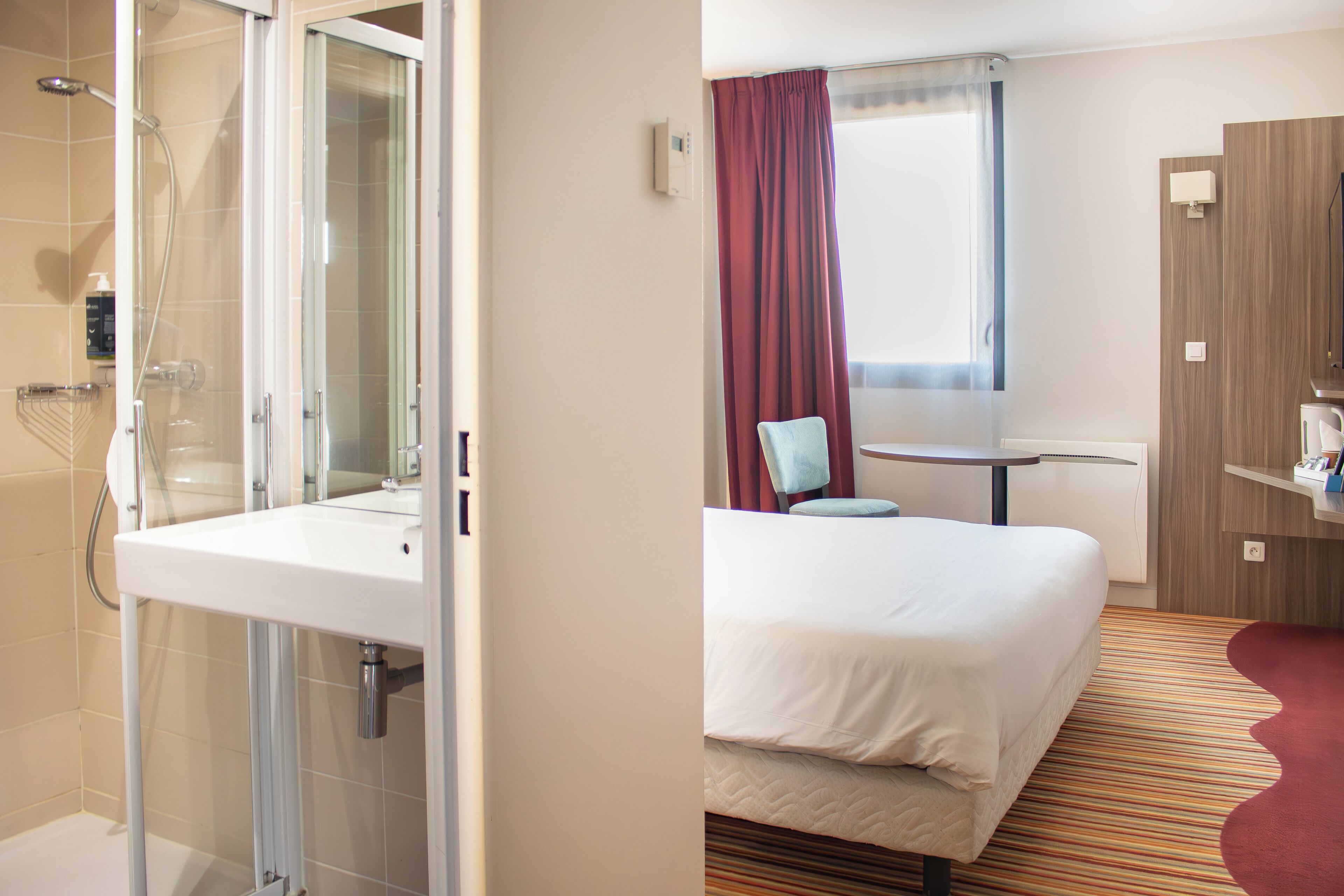 Une chambre d’hôtel avec une douche, un lit, une petite table et une fenêtre avec des rideaux.