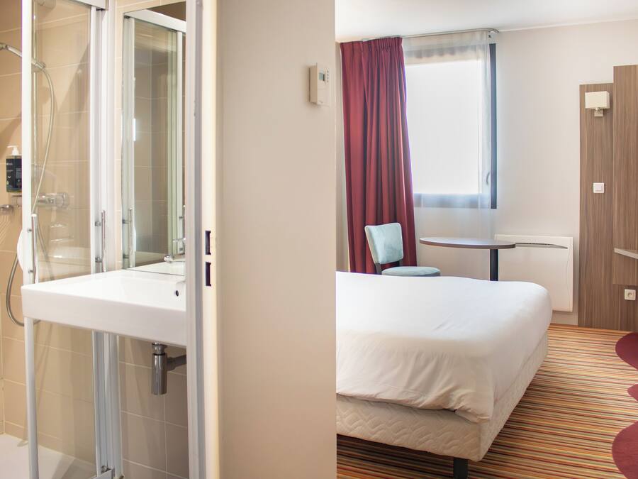 Une chambre d’hôtel avec une douche, un lit, une petite table et une fenêtre avec des rideaux.