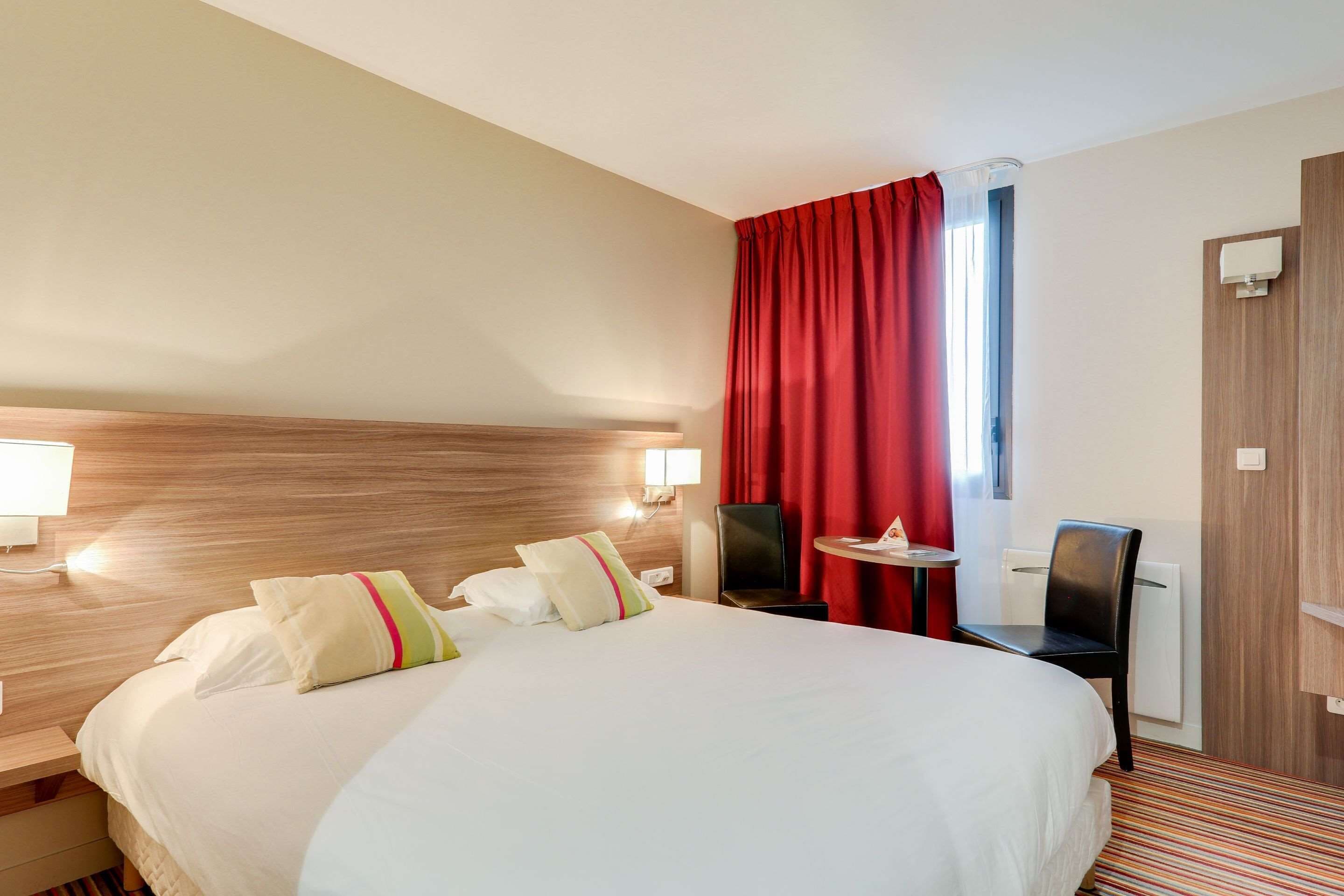 Habitación de hotel con una cama grande, dos lámparas de noche, una mesita, una silla y un ventanal con cortinas rojas.